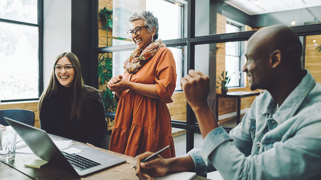 A team consisting of three people laughs in front of a laptop in an office.