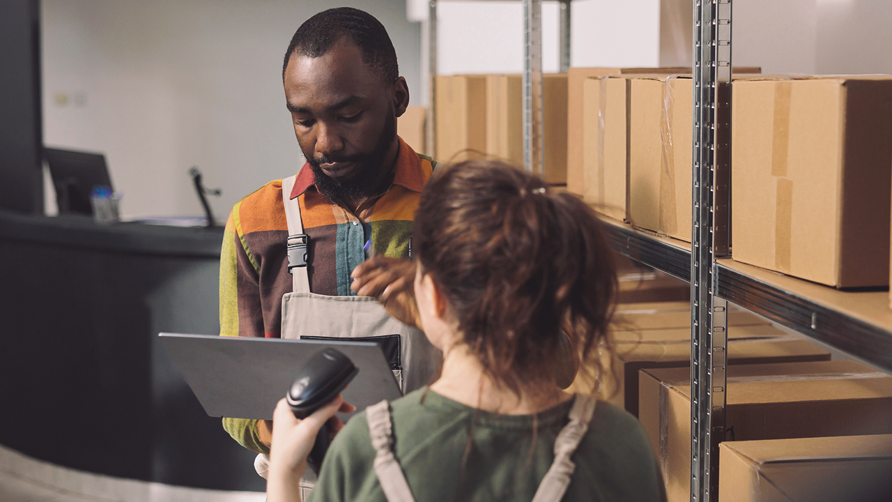 Two people in a warehouse: a woman in the foreground wearing a green top and holding a barcode scanner, a man in the background with a tablet, surrounded by shelves with boxes.