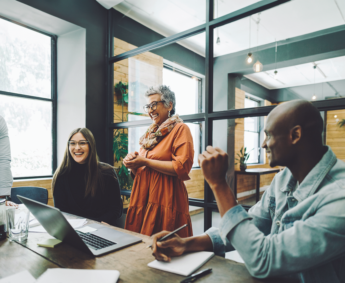 A team consisting of three people laughs in front of a laptop in an office.
