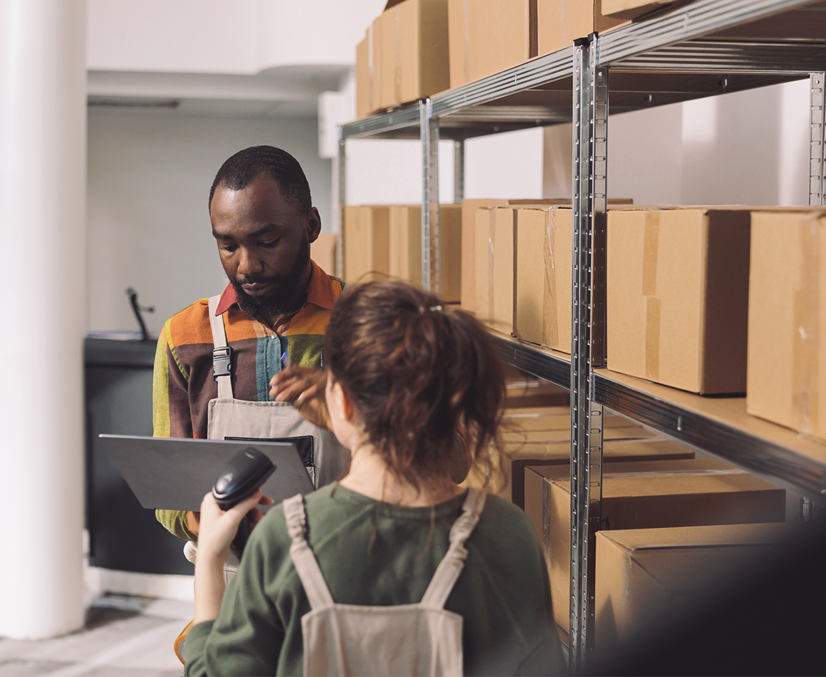 Two people in a warehouse: a woman in the foreground wearing a green top and holding a barcode scanner, a man in the background with a tablet, surrounded by shelves with boxes.