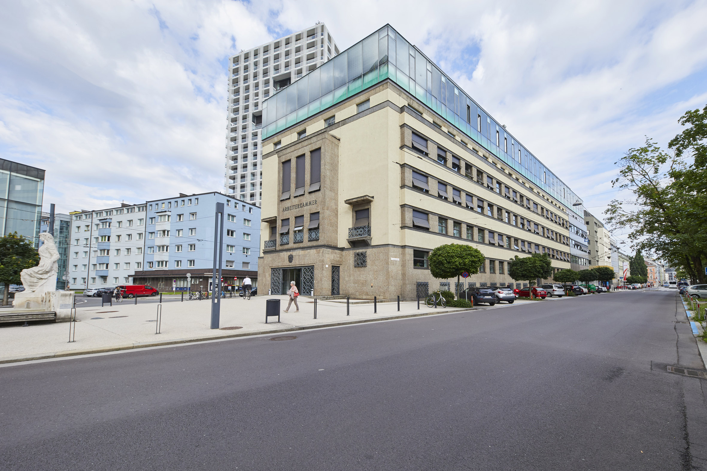 Straßenansicht des Gebäudes der Arbeiterkammer Oberösterreich mit modernen Glasanbauten, umgeben von blauen Gebäuden, Bäumen und parkenden Autos
