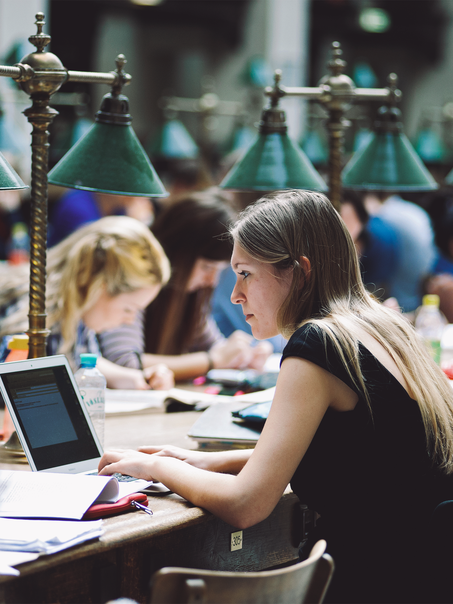 A student at the University of Vienna sits at a laptop in the library, with other students studying in the background.