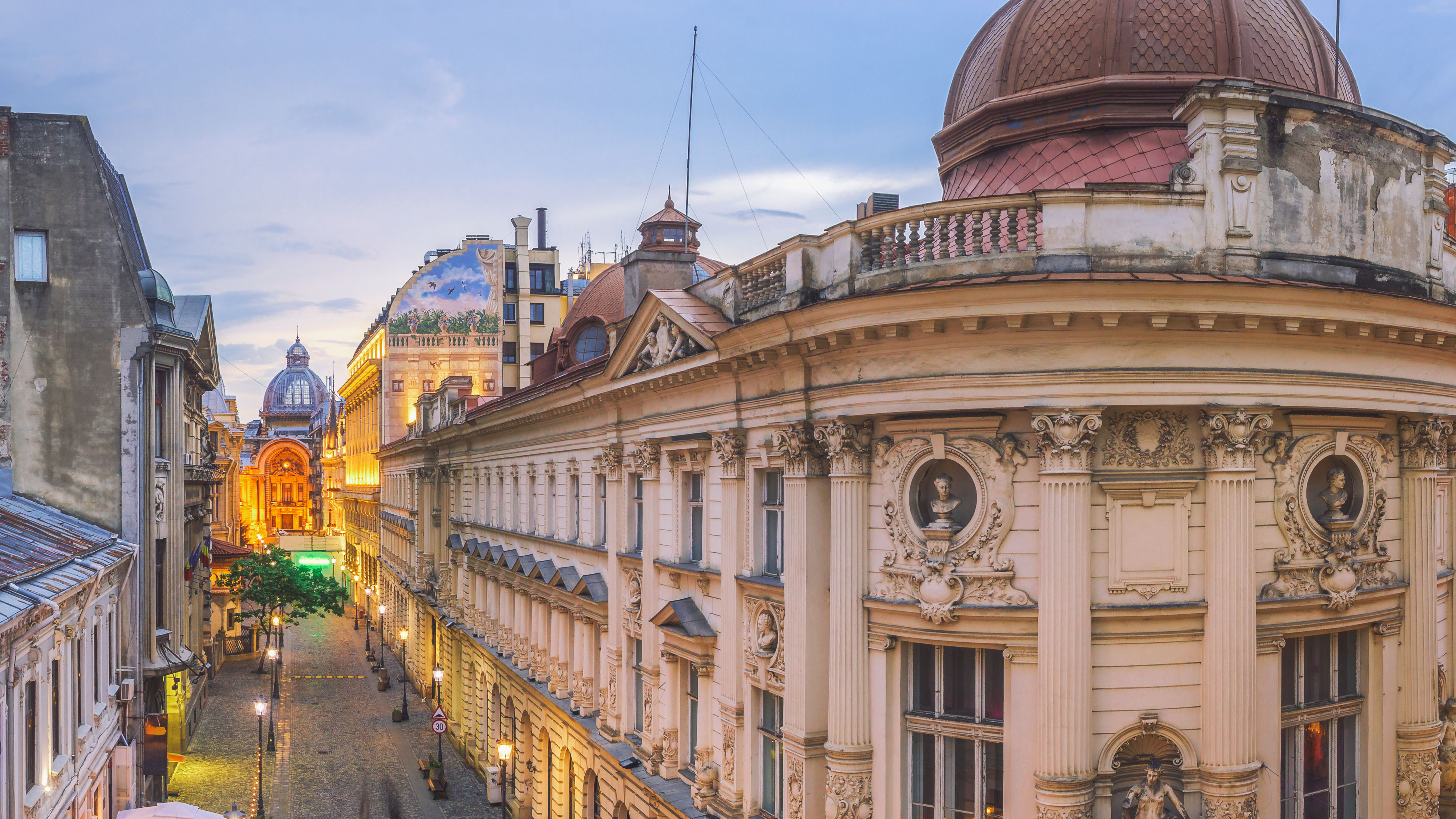 Evening view from slightly above the rooftops of Bucharest, overlooking a street in the historic Old Town with warm lighting and a calm atmosphere.