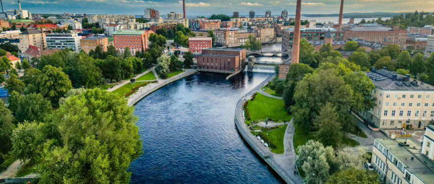 Aerial view of Tampere, Finland, showing a river flowing through the city with tree-lined banks, red-brick industrial buildings, smokestacks, and a mix of modern and historic architecture under a partly cloudy sky.