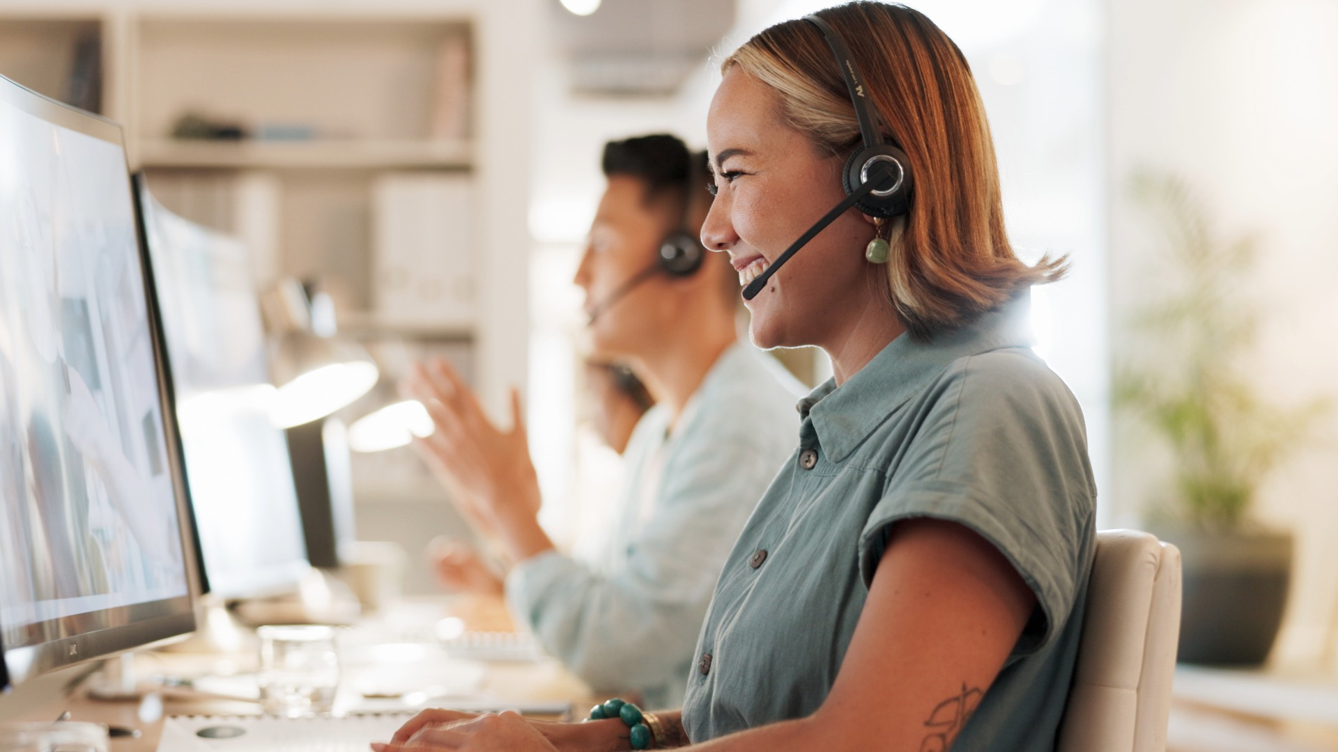 A woman is sitting in an office, smiling, wearing a headset, and working on a computer.