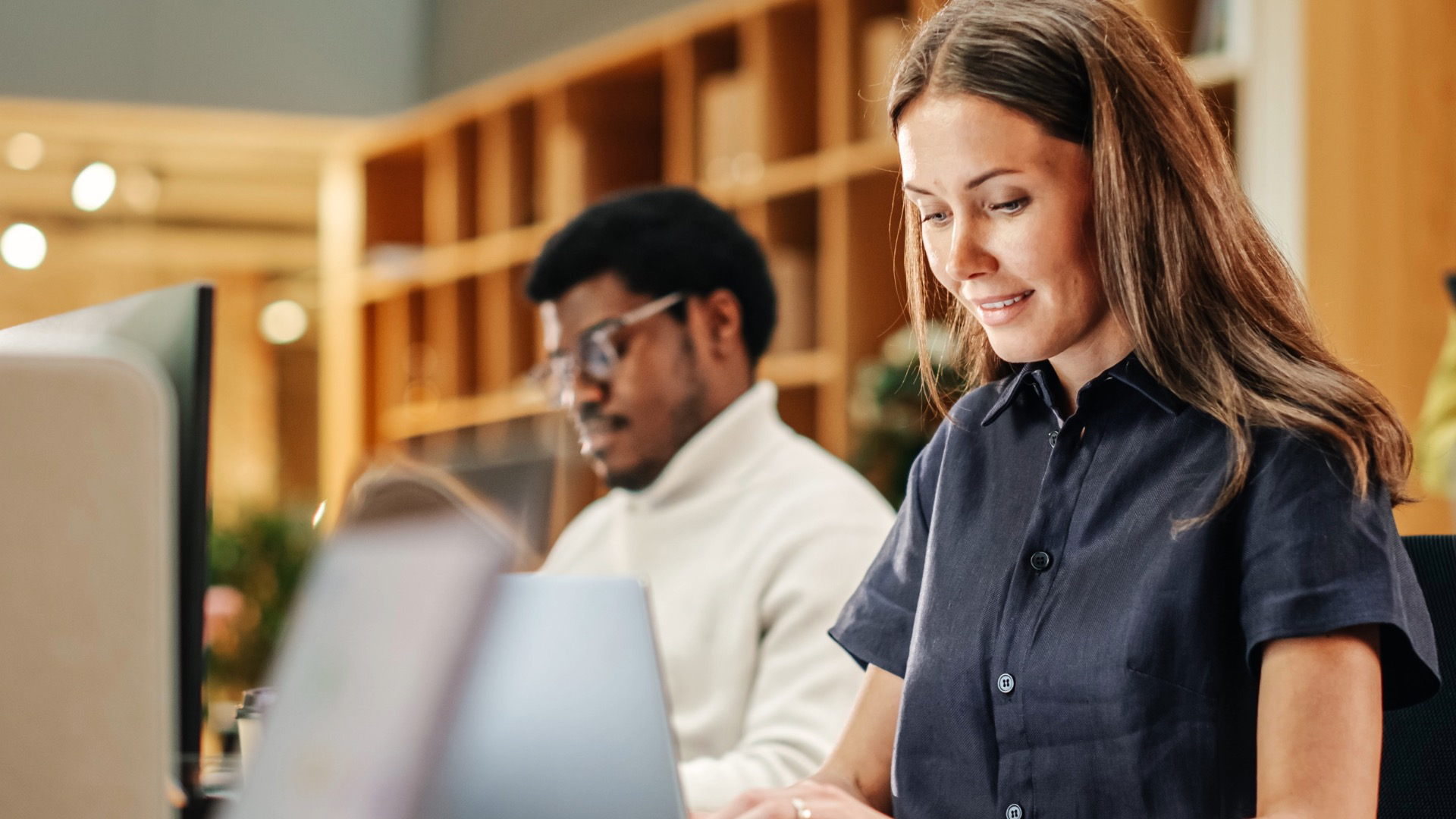 Woman and man working on laptops in an open-plan office with shelves in the background