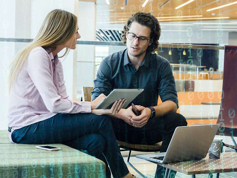 Two people are sitting opposite each other in an office, surrounded by laptops, tablets, and smartphones. In the background, there is a glass railing with binary code overlaid on it, creating a technological atmosphere.