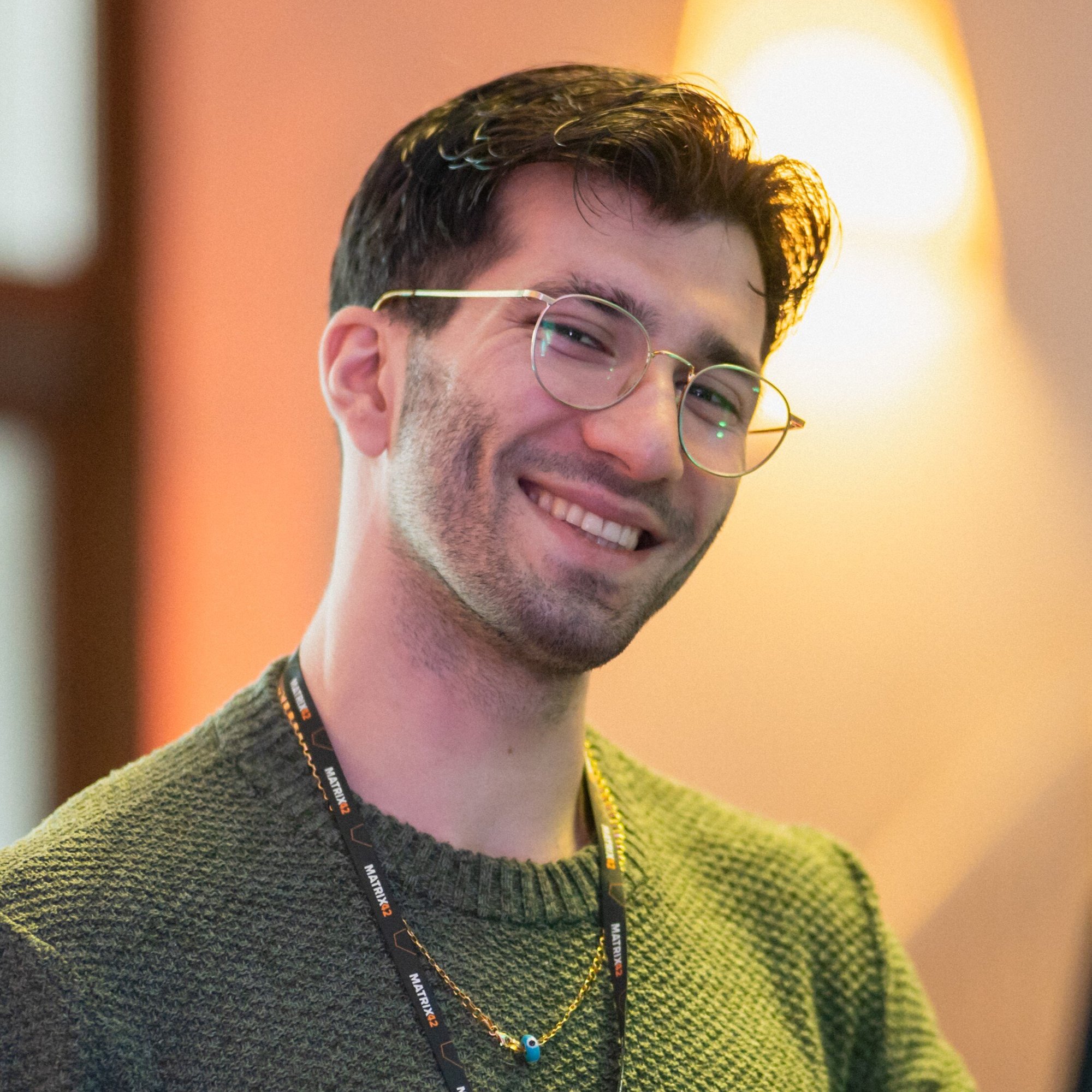 Ufuk Demirel, smiling with glasses and a green sweater, standing in warm indoor lighting