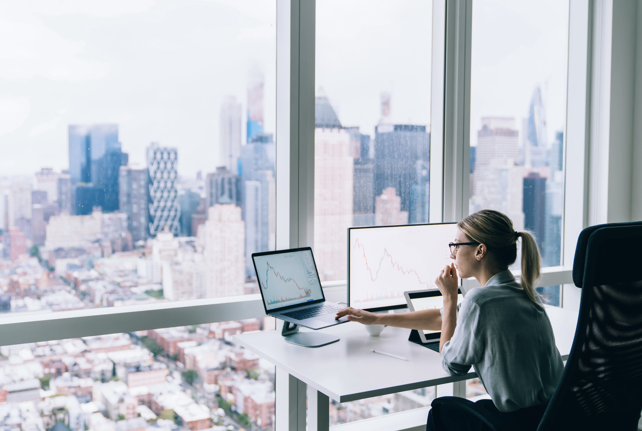 Eine Frau sitzt in einem Büro an einem Schreibtisch vor einem großen Fenster. Die Aussicht ist eine Skyline einer Großstadt