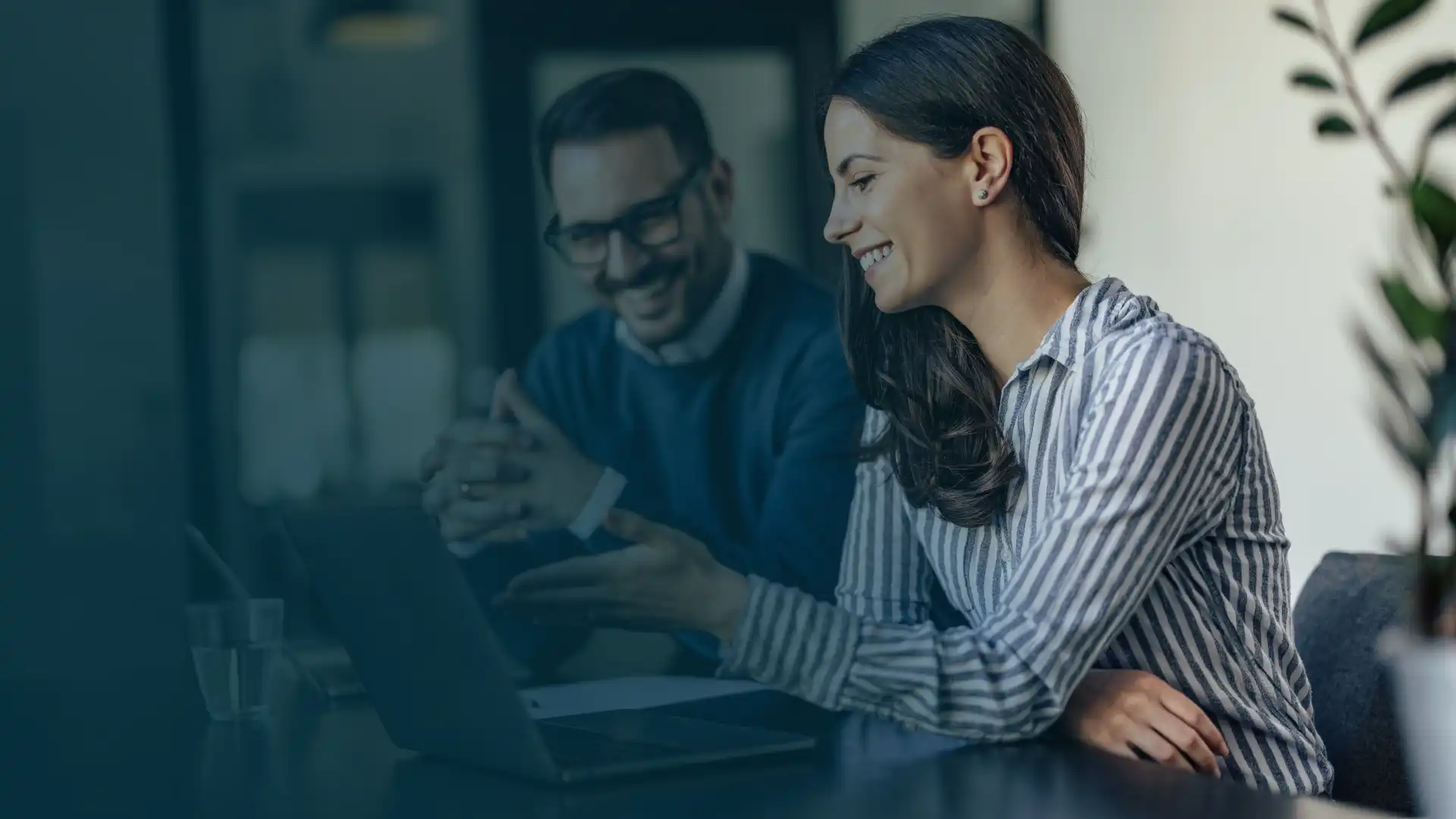 Due professionisti in una riunione di amministrazione e finanza, con una donna che gesticola seduta a un tavolo con un computer portatile e un uomo che ascolta con attenzione, in un ambiente di lavoro collaborativo.