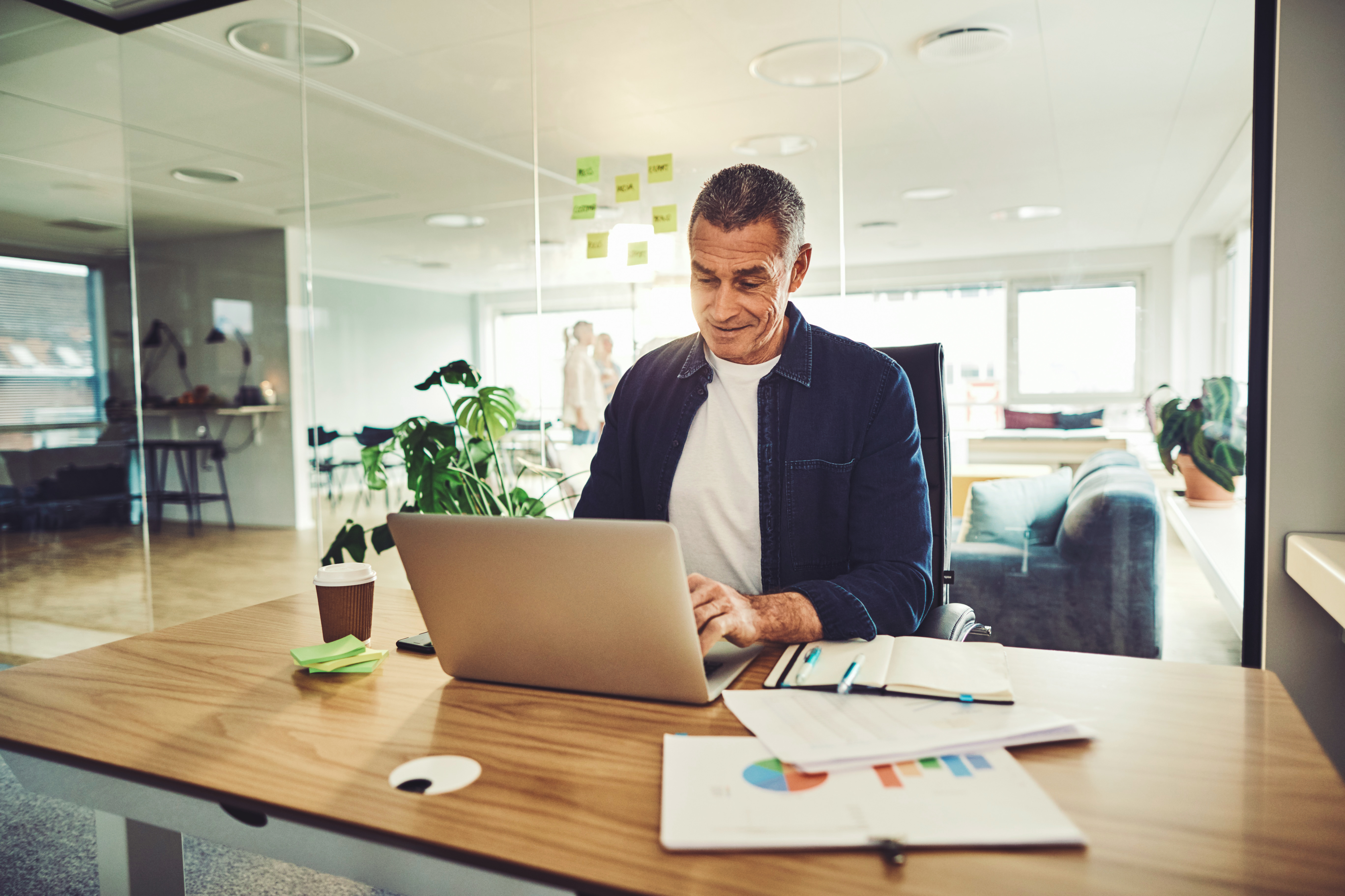 A man is working in his home office, with a laptop and papers displaying diagrams in front of him.