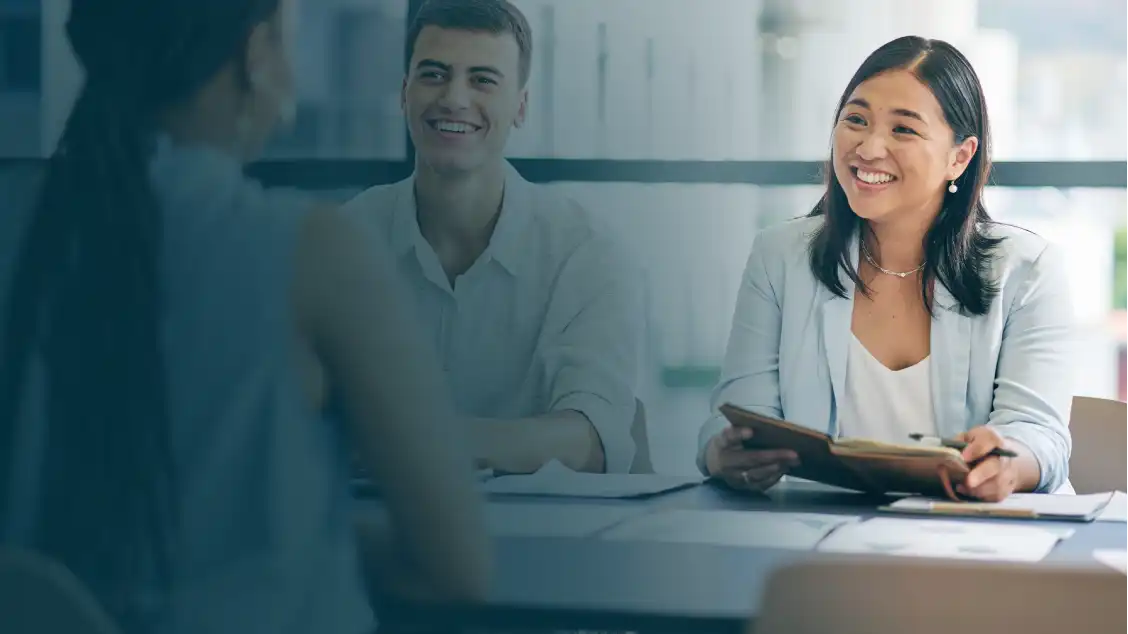 Tre persone in una riunione professionale delle risorse umane, con una donna in blazer azzurro che sorride e tiene in mano un taccuino, un uomo in camicia chiara che sorride e un'altra donna con i capelli intrecciati rivolta verso l'esterno, in un ufficio luminoso.