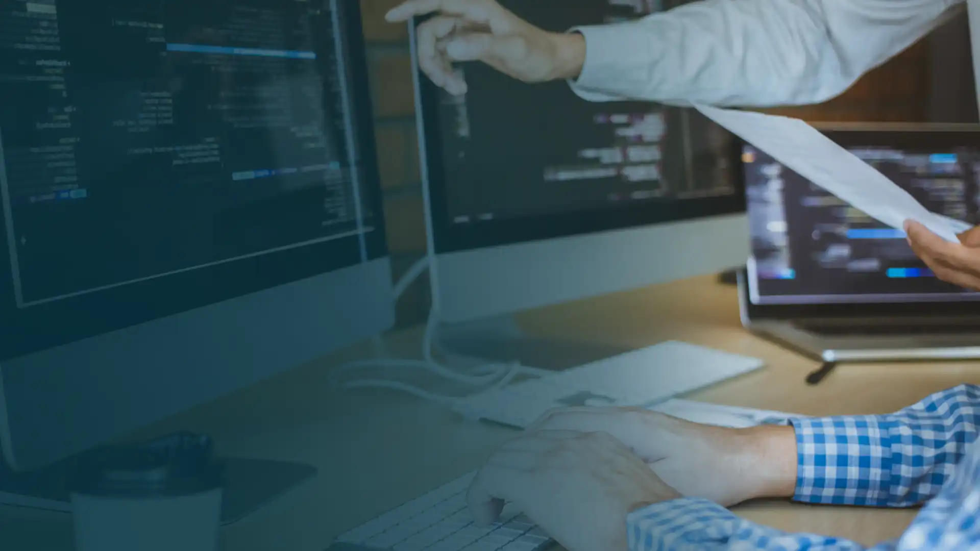 Due colleghi che collaborano alla gestione del rischio: uno indica un codice su un monitor mentre l'altro scrive su una tastiera, circondato da schermi multipli e da una tazza di caffè in un ufficio moderno.